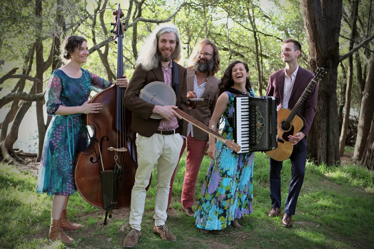 The five members of The Good Behaviours laughing under trees, holding double bass, banjo, fiddle, accordion and guitar