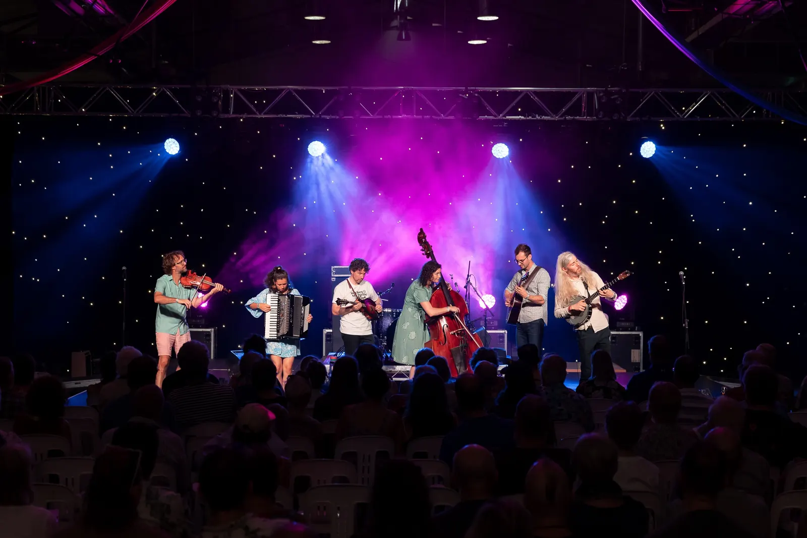 The band on a festival stage under blue and magenta lights, seen from the back of a seated audience